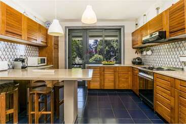 This kitchen features wooden cabinets, a tiled backsplash, modern appliances, and a large window with a garden view. Two pendant lights and a breakfast bar.