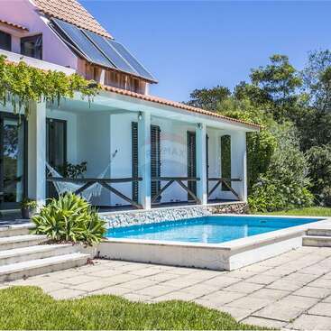Ein modernes Haus mit Sonnenkollektoren, einem Ziegeldach und einer überdachten Terrasse. Draußen gibt es eine Hängematte, üppiges Grün und einen strahlend blauen Swimmingpool.