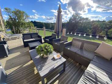 Um pátio externo aconchegante com móveis modernos, uma mesa com bebidas, luzes decorativas, plantas e uma vista incrível de árvores e um céu ensolarado e parcialmente nublado.
