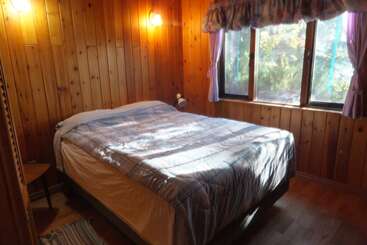 Un acogedor dormitorio con paredes de paneles de madera, una gran ventana con cortinas moradas y una cama bien hecha. Las luces cálidas crean un ambiente rústico y acogedor.