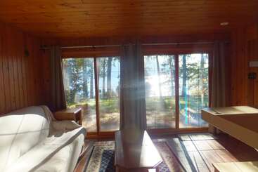 Esta acogedora sala de estar de cabaña de madera cuenta con un sofá blanco, una mesa de centro, grandes ventanales con cortinas y ofrece una serena vista de los árboles y un lago.