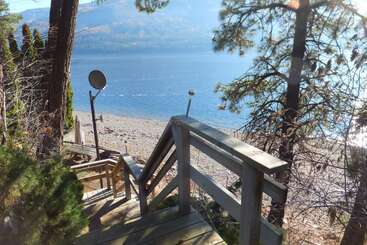 Unas escaleras de madera descienden entre altos árboles hacia una playa de guijarros junto a un lago azul, con montañas al fondo y una antena parabólica cerca.