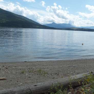 Un lac calme bordé d'une plage de galets et de rondins, avec des collines vertes et de lointaines montagnes bleues sous un ciel lumineux rempli de nuages blancs.