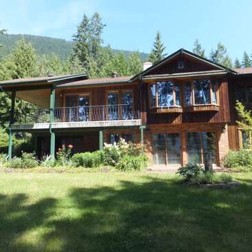 The image depicts a large, two-story house with a brown exterior and green trim, situated in a wooded area with a mountainous backdrop.