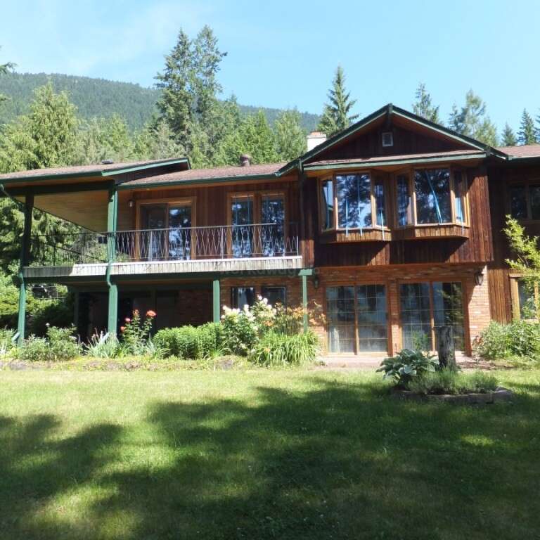 La imagen muestra una gran casa de dos plantas con el exterior marrón y ribetes verdes, situada en una zona boscosa con un fondo montañoso.