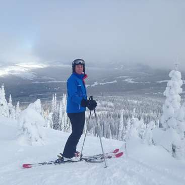 A skier stands on a snowy slope, wearing a blue jacket, black pants, and helmet, with skis and poles, set against a mountainous winter landscape.