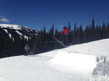 The image depicts a skier in mid-air, executing a jump on a snowy slope, with a forest of trees and a mountain in the background under a clear blue sky.