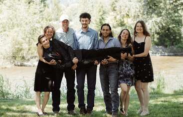 The image shows a group of people posing for a photo, with some individuals standing and others seated, all facing the camera, in front of a natural outdoor setting.