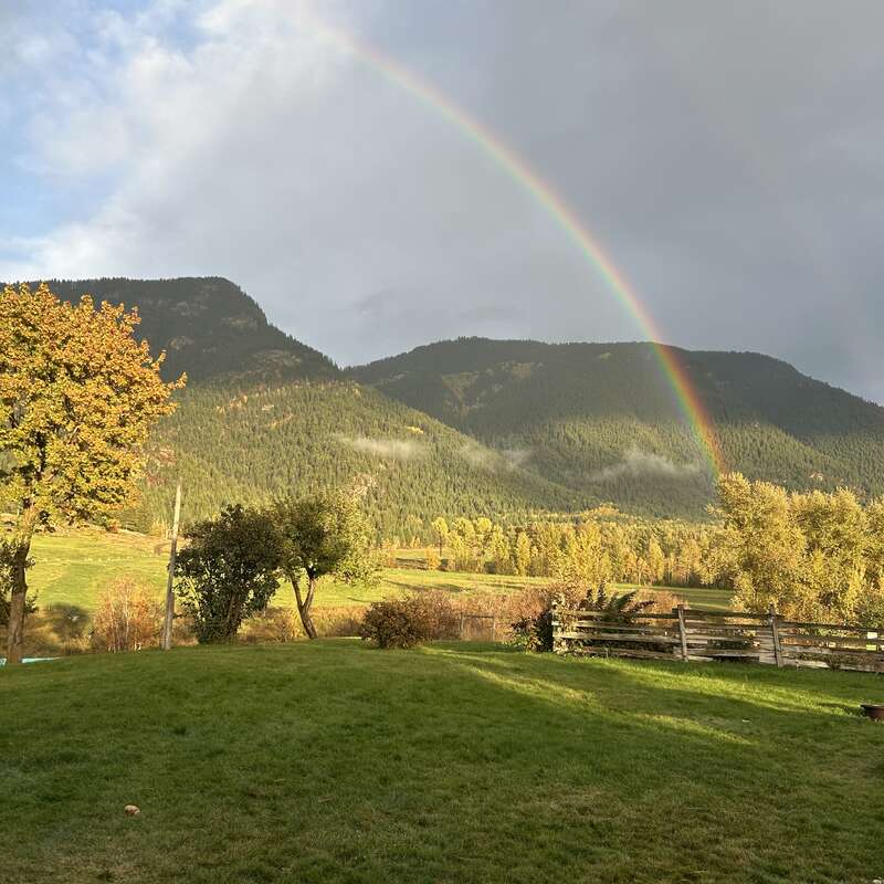 A vibrant rainbow arcs over lush green hills and forested mountains. Sunlight illuminates autumn trees, while a wooden fence and grassy field complete the tranquil scene.