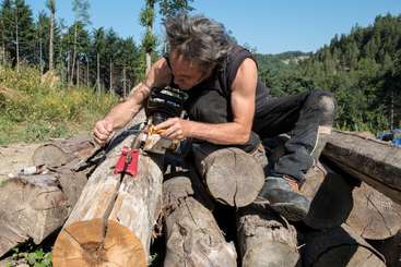 Un homme travaille avec du bois, peut-être pour construire quelque chose. Il porte un t-shirt et un short et est entouré de planches de bois et d'autres matériaux. L'arrière-plan est une zone herbeuse.