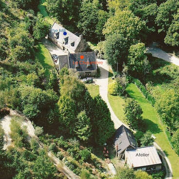 Cette image aérienne montre trois bâtiments identifiés – Le Moulin, La Chaumière et L'Atelier – nichés au milieu d'arbres luxuriants, reliés entre eux par des sentiers sinueux et des pelouses verdoyantes.