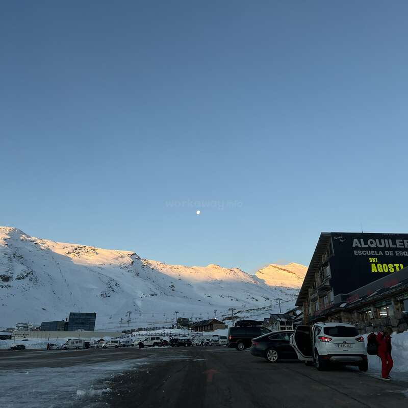 Eine verschneite Berglandschaft bei Sonnenaufgang oder Sonnenuntergang, mit Autos, die entlang einer Straße geparkt sind, einem Skiverleih und Menschen, die sich auf winterliche Aktivitäten im Freien vorbereiten.