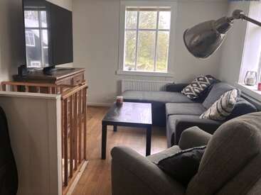 Uma sala de estar aconchegante com um sofá seccional cinza, almofadas estampadas, uma mesa de centro preta, janelas grandes, TV em um armário e piso de madeira.