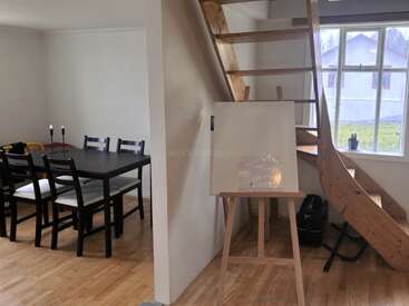 Esta imagem mostra uma sala aconchegante com piso de madeira, uma mesa de jantar preta com seis cadeiras, uma escada, uma grande janela e um cavalete de artista.