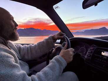 Um homem com barba dirige um carro em uma rota cênica à beira de um lago ao pôr do sol. Nuvens laranja e roxas vibrantes iluminam o céu, com montanhas em silhueta do lado de fora.