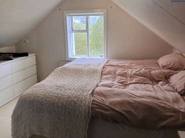 Esse quarto aconchegante no sótão tem uma cama grande com roupa de cama rosa, um cobertor texturizado, paredes brancas, uma janela com vegetação do lado de fora e uma cômoda branca elegante.