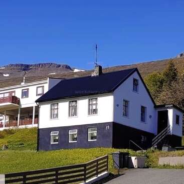 Ein charmantes weißes Haus mit dunklen Zierleisten steht auf einem grasbewachsenen Hügel. Dahinter liegen Berge mit Schneeflecken unter einem klaren blauen Himmel. Eine friedliche Umgebung.