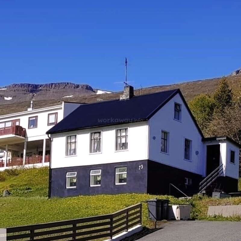 Uma charmosa casa branca com acabamento escuro fica em uma colina gramada. Atrás dela, há montanhas com manchas de neve sob um céu azul claro. Um cenário tranquilo.