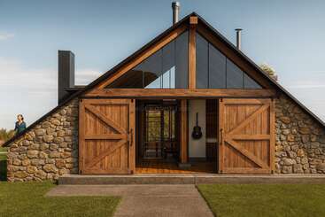 Eine rustikale Hütte aus Stein und Holz mit großen Scheunentoren, dreieckigen Fenstern und einer Gitarre, die im Inneren hängt. Auf der linken Seite steht eine Person, die die Natur genießt.