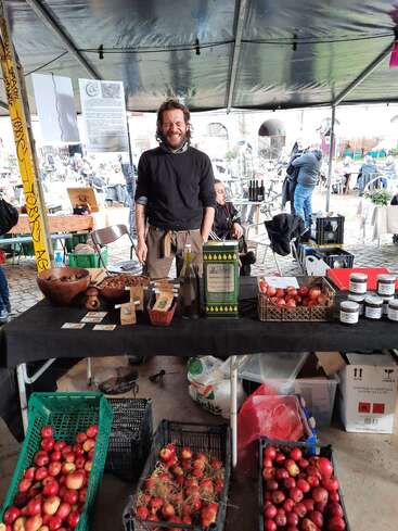 Das Bild zeigt einen Mann, der hinter einem Tisch auf einem Markt im Freien steht, umgeben von verschiedenen Lebensmitteln und Produkten, mit einer Menschenmenge im Hintergrund.