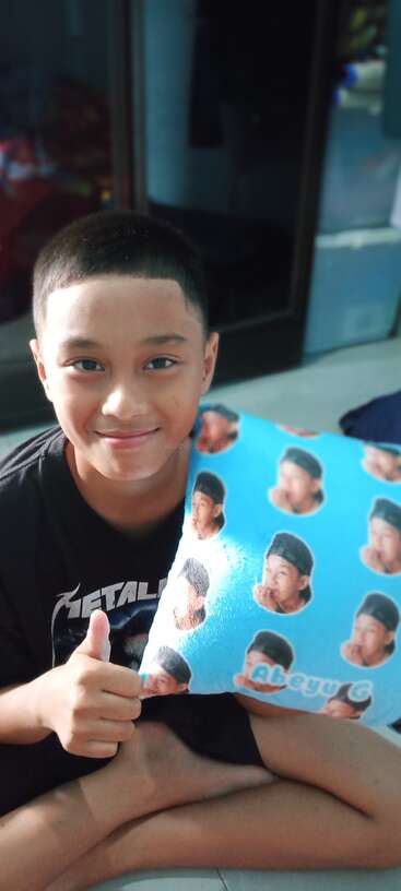 A young boy smiles while sitting on the floor, holding a blue pillow printed with faces and wearing a black Metallica shirt, giving a thumbs-up.