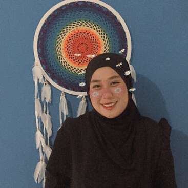 A smiling woman in a black hijab stands in front of a blue wall, with a colorful dreamcatcher. Filter adds daisy stickers to her cheeks and head.