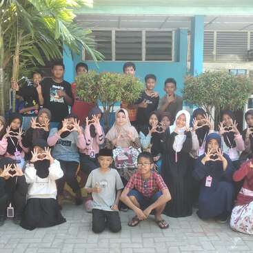 A group of children and adults poses outside a building, many making heart shapes with their hands, smiling in a cheerful, sunny, and friendly atmosphere.