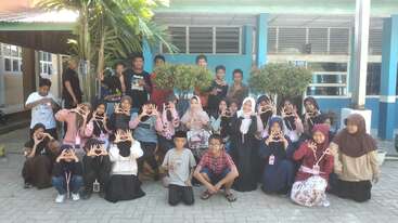 A group of children and adults poses outside a building, many making heart shapes with their hands, smiling in a cheerful, sunny, and friendly atmosphere.