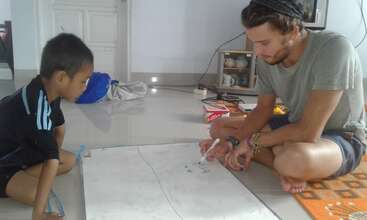 The image shows a man and a child drawing on a large piece of paper on the floor, with the child lying down and the man sitting beside him.