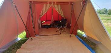 Dentro de uma barraca espaçosa, uma cadeira e uma mesa de acampamento estão dispostas sobre um tapete. A luz do sol entra pela abertura, e dá para ver a grama lá fora, criando um ambiente aconchegante e convidativo.