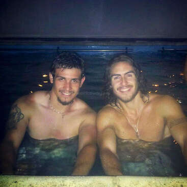 Dois rapazes sorridentes relaxam na piscina à noite. Os dois estão sem camisa, encostados na beira da piscina, claramente curtindo o momento e a amizade um do outro.