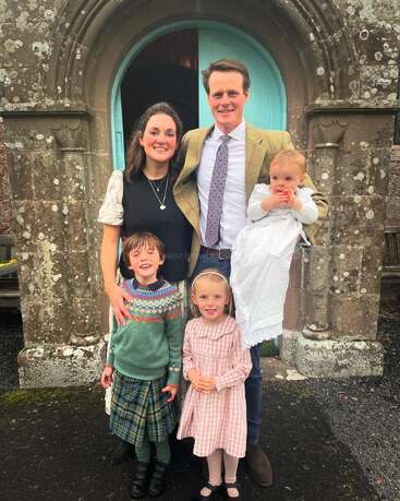 The image depicts a family of five posing for a photo in front of an old stone building with a teal door, dressed in formal attire.