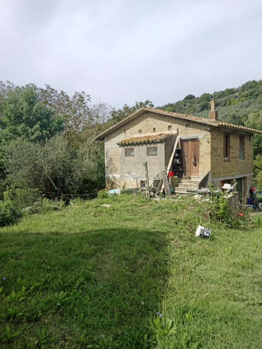 Uma pequena casa rústica fica sozinha em uma paisagem verde e exuberante. Cercado por árvores e colinas, o quintal é desarrumado, evocando uma atmosfera pacífica e rural.