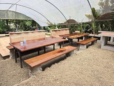 The image shows polished wooden tables and benches under a transparent canopy outdoors, surrounded by greenery and gravel ground, creating a rustic, natural, and inviting seating area.