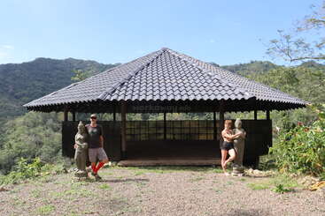 A large, open pavilion with a tiled roof stands on a hilltop, surrounded by greenery and mountains. Two people pose beside statues at the entrance.