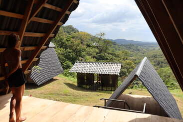 A person stands on a wooden deck, under a slanted roof, overlooking several cabins in a lush, green mountainous landscape beneath a partly cloudy sky.