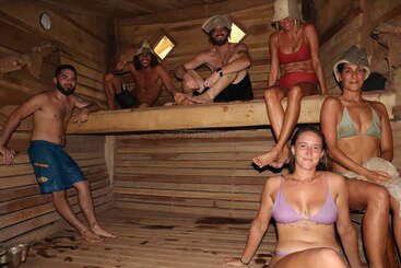 Six people relax inside a wooden sauna, wearing swimsuits and felt hats. They are smiling and seated on different levels, enjoying the warm, cozy atmosphere.