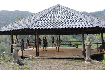 A group of people gathers under a large, open, tiled-roof pavilion in a lush, mountainous setting, flanked by two statues, for outdoor activities.