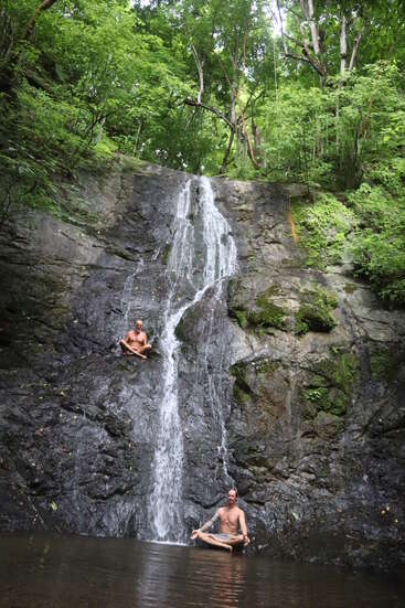 Two men meditate at a small waterfall in a lush green forest. One sits on the rocks, the other in water below, both peaceful and relaxed.