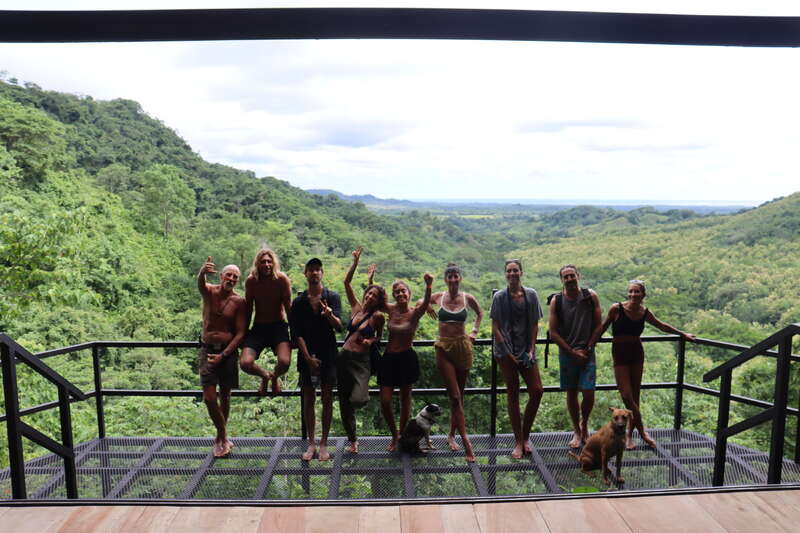 A group of ten people and two dogs pose on a metal balcony, overlooking a lush green forest and distant hills under a cloudy sky, radiating joy.
