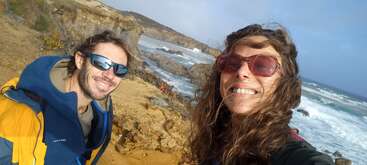 Zwei Menschen mit Sonnenbrillen lächeln für ein Selfie auf einer felsigen Klippe am Meer. Hinter ihnen rauschen die Wellen des Meeres unter einem teilweise bewölkten blauen Himmel.