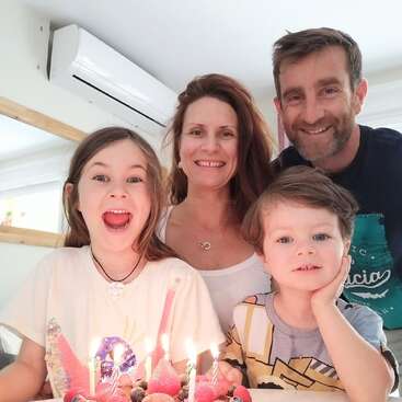 La imagen muestra a una familia de cuatro miembros posando detrás de una tarta de cumpleaños con velas encendidas, sobre un fondo blanco de pared y techo.