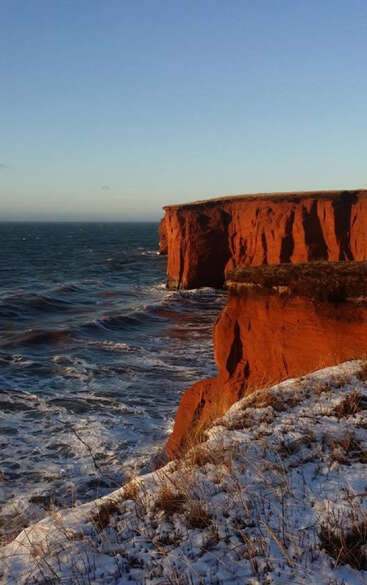 Enormes acantilados rojizos se asoman a un mar azul agitado; la hierba parcialmente nevada bordea los acantilados bajo un cielo despejado, en el que la luz del sol proyecta sombras espectaculares.