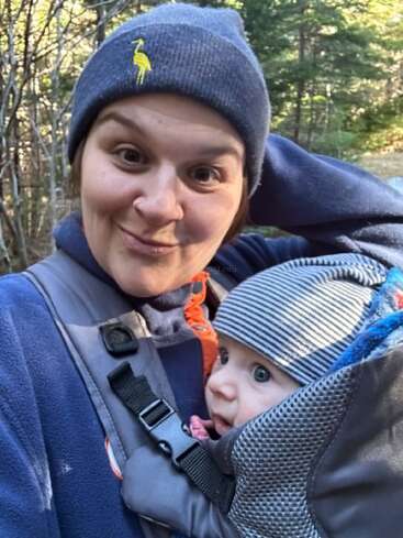 Uma mulher usando um gorro azul e uma jaqueta tira uma selfie divertida ao ar livre, carregando um bebê de olhos arregalados em um carrinho, ambos bem agasalhados em meio à natureza.