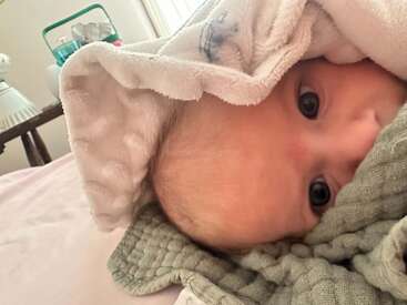 Um bebê de olhos grandes e brilhantes está aconchegado em cobertores macios. O bebê parece estar bem confortável, espreitando por baixo de uma toalha branca e fofa.