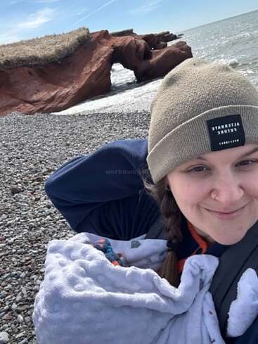 Uma mulher sorridente, com um piercing no nariz e uma trança, usando um gorro bege, segura um bebê enrolado em um cobertor numa praia rochosa, com formações rochosas vermelhas e o oceano ao fundo.