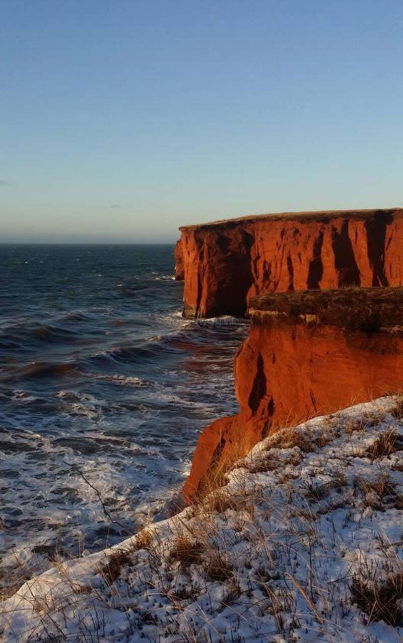 Towering red cliffs overlook a choppy blue sea, partially snow-dusted grass edges the cliffs under a clear sky, with sunlight casting dramatic shadows.