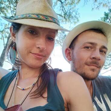 Una mujer y un hombre posan juntos al aire libre. Ambos llevan sombreros y ropa informal, y sonríen amablemente. Al fondo se ven la luz del sol y los árboles.