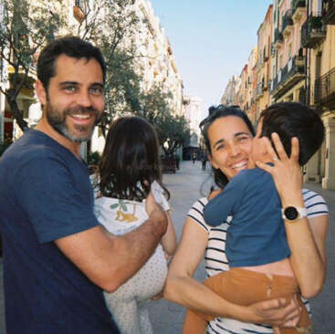 Eine glückliche Familie steht auf einer sonnigen Straße, die Eltern lächeln und halten zwei kleine Kinder im Arm. Im Hintergrund sieht man auf beiden Seiten Bäume und bunte Gebäude.