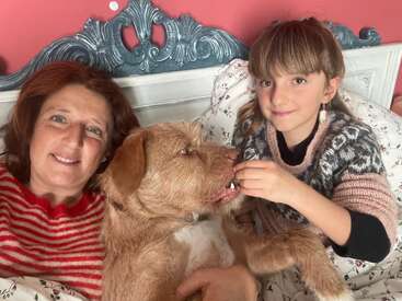 A woman, a young girl, and a brown dog relax together on a bed. They are smiling, sharing a warm, cozy, and affectionate moment.